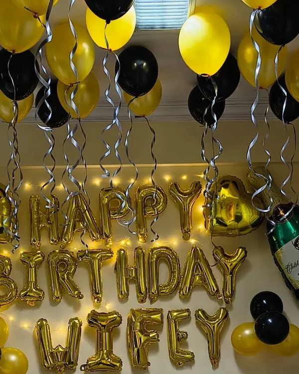 A classic birthday surprise for a loved one. This room decoration features gold and black balloons on the ceiling, "Happy Birthday Wifey" foil letters, and champagne bottle foils for a celebratory touch.