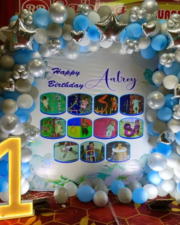 An adorable elephant-themed first birthday party backdrop. The setup features a balloon arch in blue, silver, and white, a large light-up number '1', and a personalized backdrop with the baby's monthly milestone photos.
