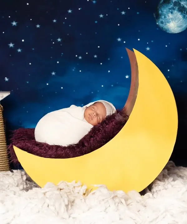 A peaceful newborn, swaddled in white, rests on a bright yellow crescent moon against a magical starry night sky. We created this dreamy celestial theme in our Jaipur studio to capture these fleeting, precious moments.