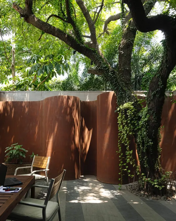 An outdoor seating area at the KEUS experiential hub, nestled against a curving Corten steel wall and shaded by a mature tree. This space is designed as a breakout zone for work or relaxation, blending industrial materials with natural elements.