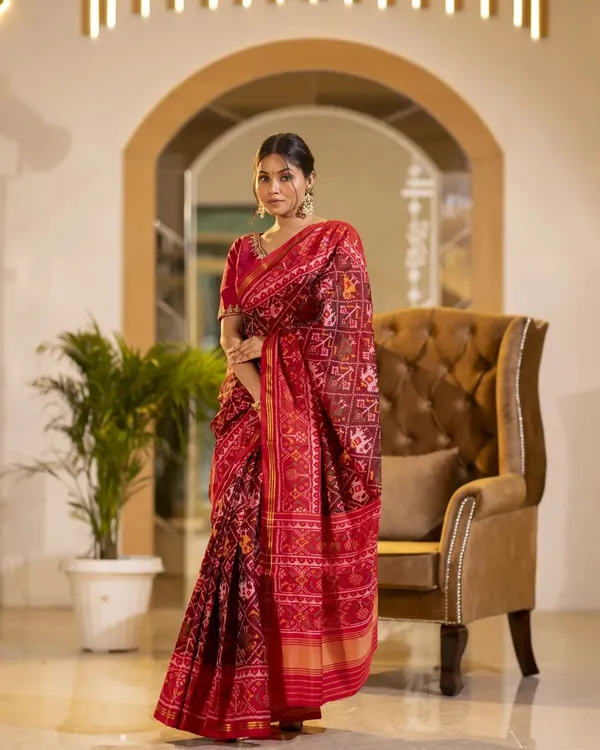 A full-length shot of a model in a traditional red Patola saree. The elegant standing pose in a well-appointed room highlights the drape and richness of the fabric, perfect for festive wear collections.