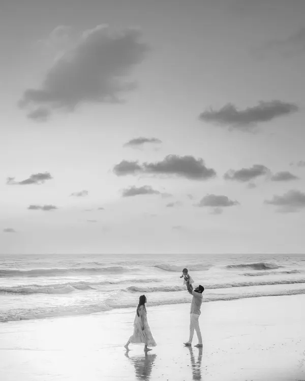 A striking black and white photo of a family on the beach. The father joyfully lifts their child into the air while the mother walks nearby, creating a dynamic and artistic family portrait.