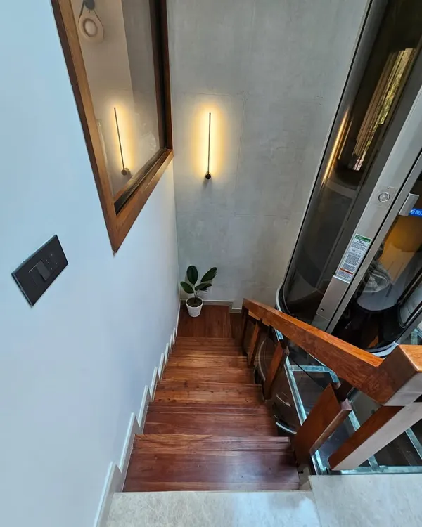A top-down view of the stairwell, showcasing the sleek, all-glass vacuum elevator alongside Indonesian walnut stair treads. Ambient wall lighting and a touch of greenery complete this statement of luxury and smart living.