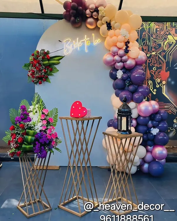 A stylish "Bride to be" setup with a modern, edgy vibe. The decor features a white backdrop with a neon sign, an asymmetrical balloon arch in rich tones, and fresh floral arrangements on gold stands.