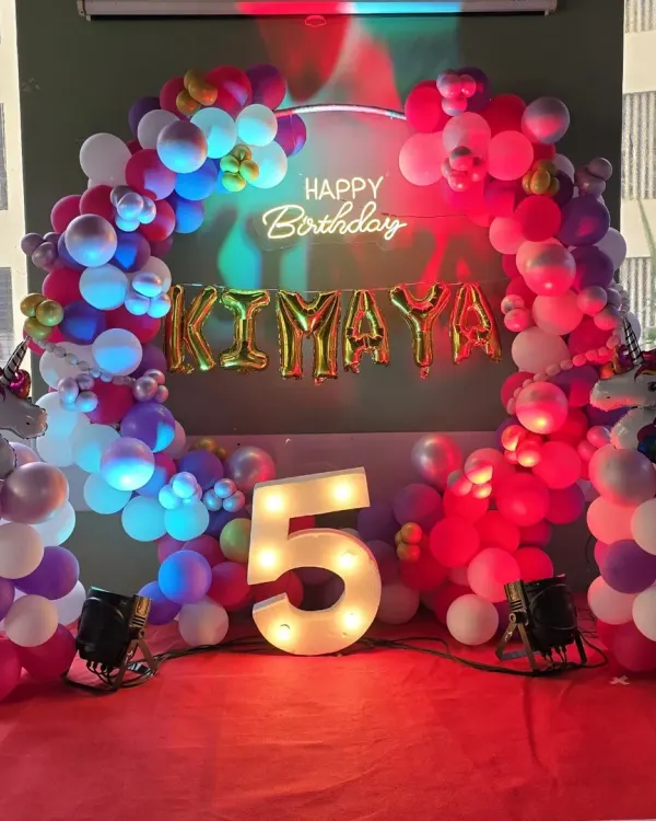 I create magical unicorn wonderlands like this one for a special 5th birthday. This setup features a vibrant balloon arch on a ring frame, a custom 'Happy Birthday' neon sign, and a large marquee number '5' to mark the milestone.