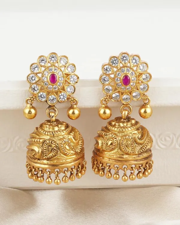 Classic gold-toned jhumkas featuring a floral stud set with white stones and a central ruby-colored gem. The bell is intricately carved, offering a timeless and versatile design.