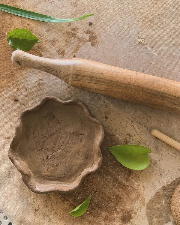 A hand-built bowl with a leaf impression, sitting next to a wooden rolling pin. In its raw, unfired state, you can see the delicate details and the soft texture of the natural clay.