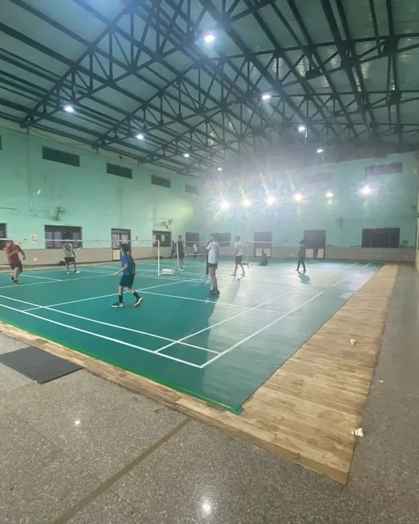 This is our indoor badminton facility at Pitampura, featuring multiple green synthetic mat courts. The space is well-lit for evening and late-night games, with ample room between courts for players.
