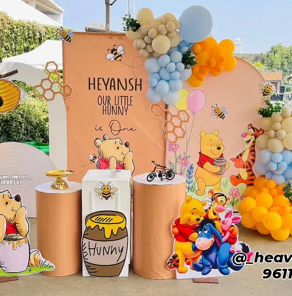 A delightful Winnie the Pooh "Our Little Hunny" first birthday party. This outdoor setup features character cutouts, honey-pot-themed plinths, and a large number '1' filled with balloons in a pastel color scheme.