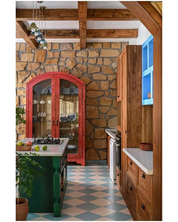 This kitchen is designed to feel like it's from another time, miles away from fleeting trends. We used a bold color palette with a green island, a red crockery cabinet, and a checkered floor, all set against a rugged stone wall and exposed wooden beams. Every element is chosen to create a warm, rustic, and deeply personal cooking space.