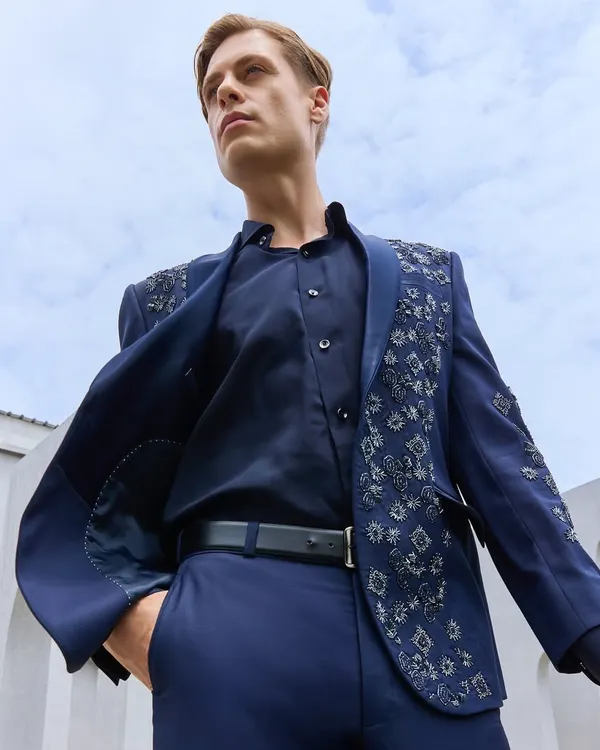 A powerful look for a modern gentleman. This deep navy suit jacket features handcrafted silver embroidery on the shoulders and lapel, offering a bold yet sophisticated statement for any reception or cocktail event. The half-lined construction ensures a comfortable, flattering silhouette.