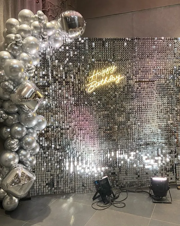 This modern and chic birthday setup features a full silver sequin shimmer wall, creating a dazzling photo backdrop. It's complemented by a silver chrome balloon garland and a warm 'Happy Birthday' neon sign, perfect for an elegant adult celebration.