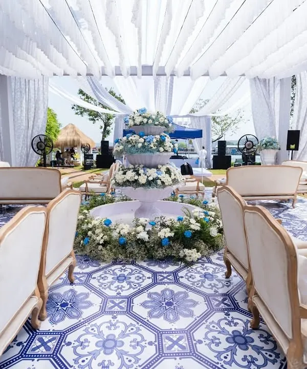 A wide view of a French-inspired carnival, with a central floral fountain and lounge seating arranged on a custom blue and white tiled floor. The entire setup is under a white canopy, creating a bright and airy feel.