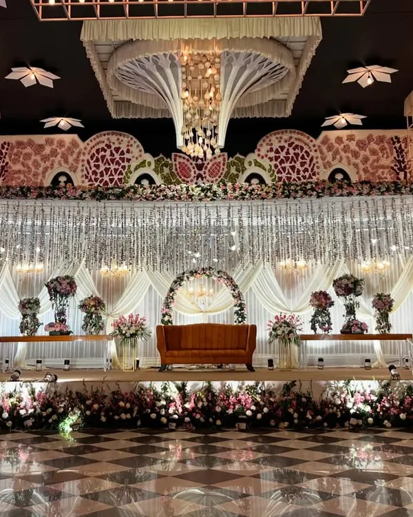 A breathtakingly grand reception stage. The design features a massive ceiling installation, a backdrop of hanging crystals, and a multi-tiered stage filled with pink and white floral arrangements.