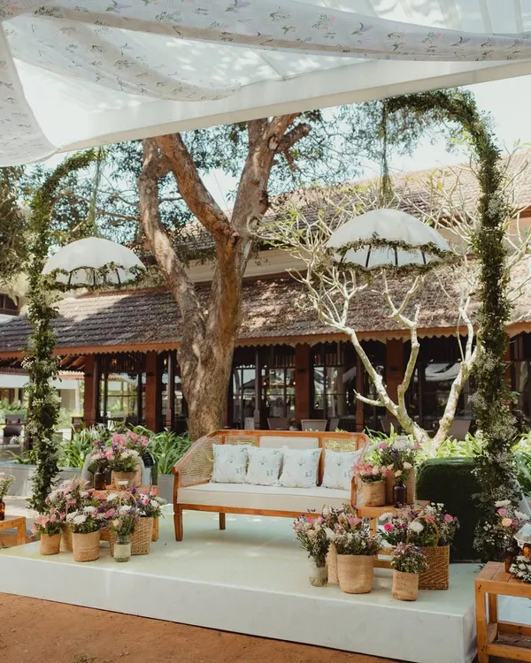 A minimalist Mehendi stage designed for a laid-back Goan wedding. A simple wooden bench is framed by two delicate floral arches and whimsical hanging umbrellas, creating a look that is both modern and dreamy.