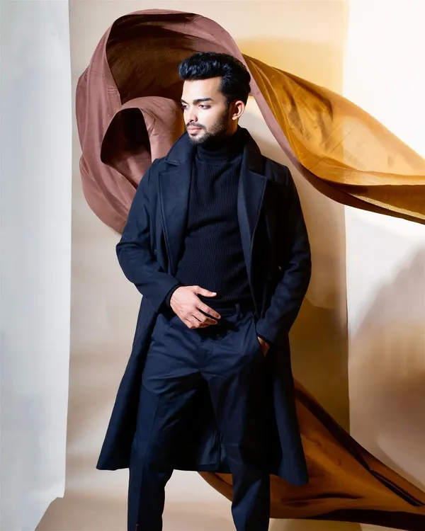 A high-fashion menswear shot featuring a model in a dark navy trench coat against an artistic, draped fabric background. This image combines classic apparel with creative studio lighting and styling.
