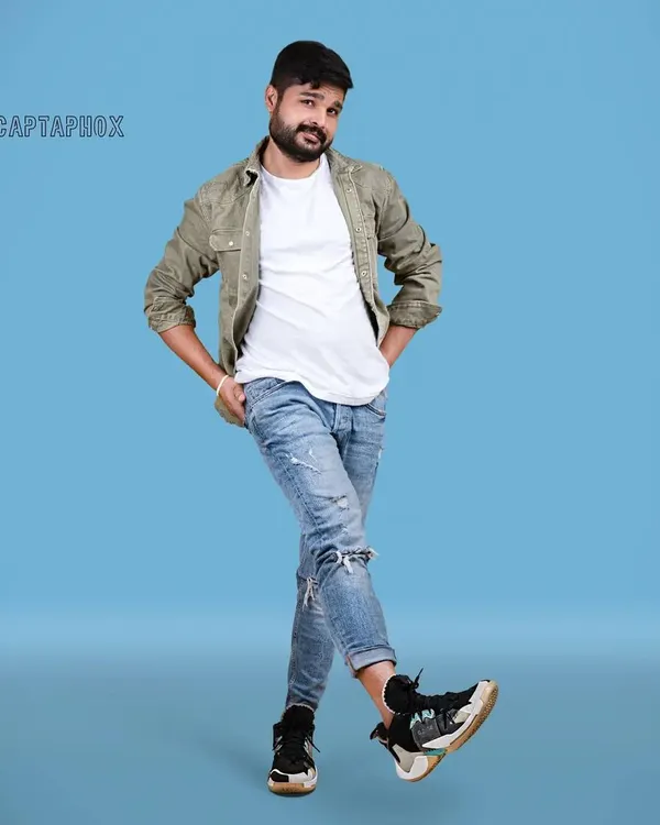 A full-length shot for a male model's portfolio, showcasing a casual, layered look. The solid blue backdrop provides a vibrant, clean setting that makes the subject and his relaxed pose the central focus.