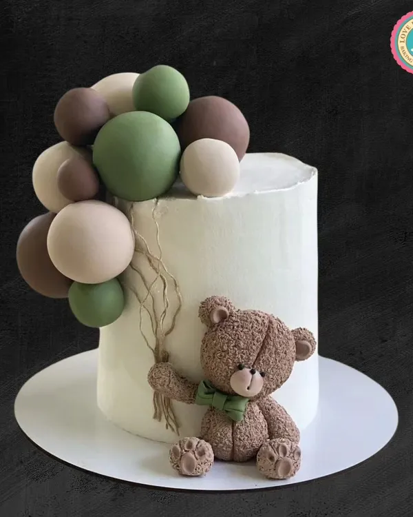 A charming teddy bear cake with a modern, earthy feel. This design features a sweet, textured teddy bear holding a bundle of green, brown, and cream-colored balloons against a clean white cake.
