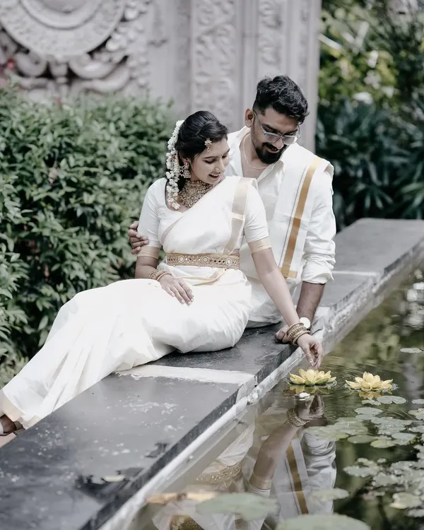 A serene and romantic pre-wedding photo of a couple in traditional white South Indian attire, sitting by a lily pond, capturing a moment of quiet intimacy.