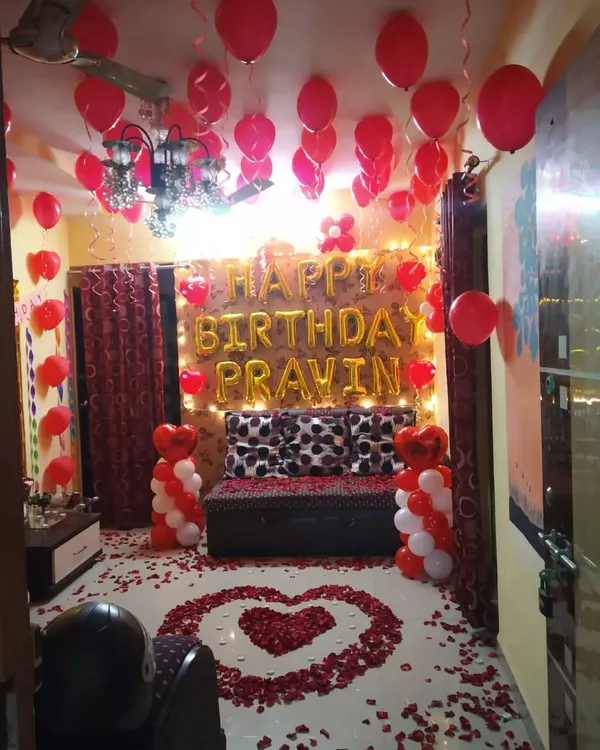 A complete romantic birthday surprise package. This room decoration includes red and white balloons, fairy lights, 'Happy Birthday' foil letters, and a heart-shaped rose petal arrangement on the floor.