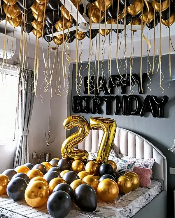 A stylish 27th birthday room decoration with black and gold balloons covering the bed and ceiling. The setup includes large number '27' foil balloons and 'Happy Birthday' letters on the wall.