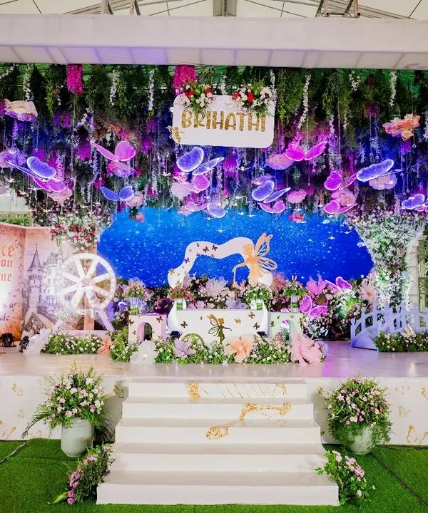 A breathtaking enchanted garden created for a darling's first birthday. This grand stage setup features a lush floral canopy with illuminated butterflies, a sparkling backdrop, and a whimsical bridge, transforming the venue into a real-life fairytale.
