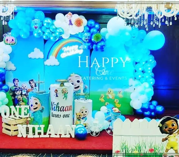 A complete Cocomelon themed first birthday party setup. The vibrant blue backdrop features the Cocomelon characters and a rainbow, framed by blue and white balloon garlands and character cutouts for an immersive experience.