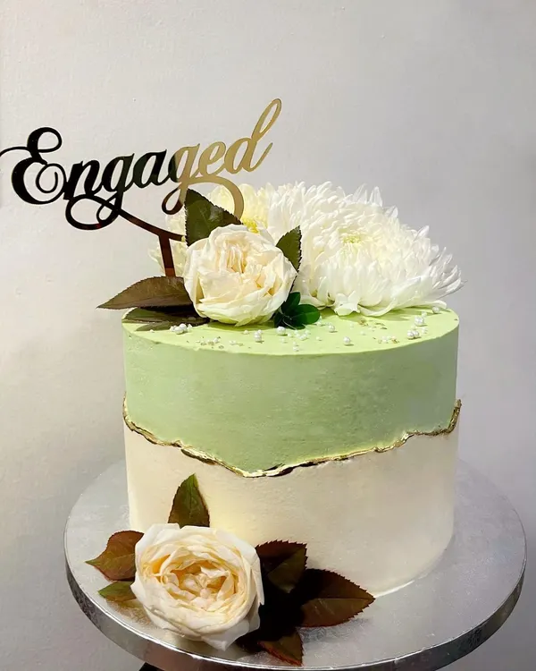 This fault-line cake was designed for an engagement party. It features a soft pistachio green top section over an off-white base, with the "break" highlighted by a delicate gold leaf edge. The design is completed with an "Engaged" topper and fresh white flowers.