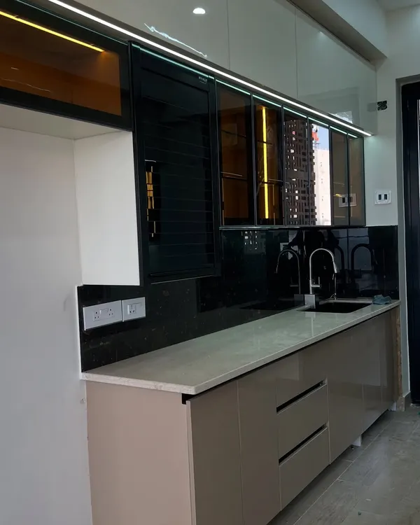 A modern kitchen featuring a mix of beige and white high-gloss cabinets, a black granite backsplash, and tinted glass upper units with profile lighting.