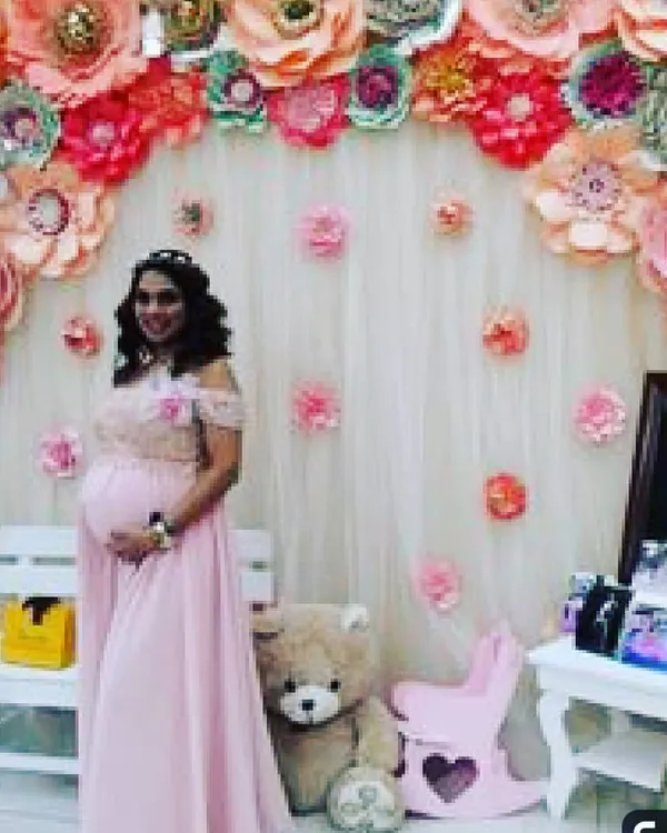A beautiful baby shower setup featuring a mother-to-be. The backdrop is a custom-made arch of handmade paper flowers in soft pinks and peaches, creating an elegant photo corner. Props like the teddy bear and rocking horse add a charming touch to the scene.