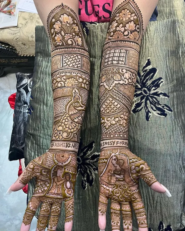 A stunning example of full bridal portrait mehndi. This design includes bride and groom figures, the wedding hashtag, and peacock motifs, all woven together with dense, traditional patterns that cover the hands and arms completely.