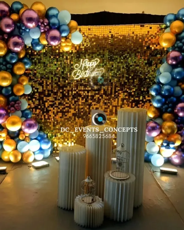 A glamorous birthday photo booth featuring a gold shimmer wall framed by a colorful balloon arch. The setup is completed with decorative pedestals and a custom neon "Happy Birthday" sign.