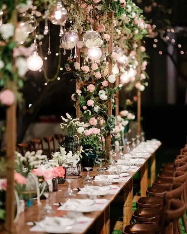 An enchanting long table setup for an evening reception, with a wooden structure overhead covered in flowers and hanging lights.