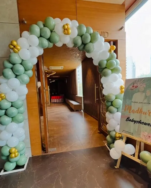 A lovely balloon arch in sage green and white for a baby shower entrance, complemented by a personalized welcome sign on an easel.