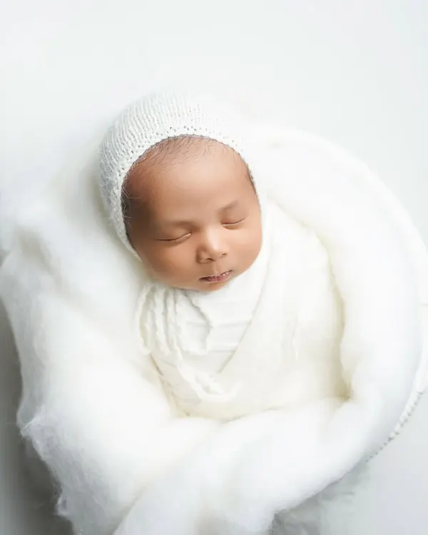 An 18-day-old baby boy, swaddled in soft white wool and a delicate bonnet. This portrait highlights the serene and pure essence of a newborn, captured with a minimalist approach.