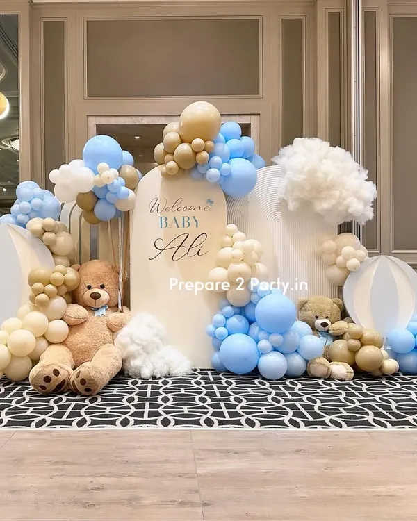A luxurious and dreamy welcome home setup for "Baby Ali" in a grand hall. The design features soft blue, beige, and white balloons creating a cloud-like effect around arched backdrops, complete with large teddy bears and orb props.