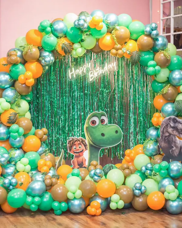 A roaring good time with a dinosaur-themed birthday! This setup features a vibrant green and orange balloon arch, a neon sign, and friendly dinosaur character cutouts.