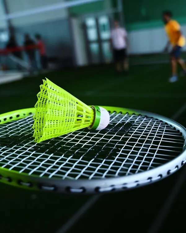 A close-up of a shuttlecock resting on a badminton racket, with players rallying in the background. It's the small details that make the game.
