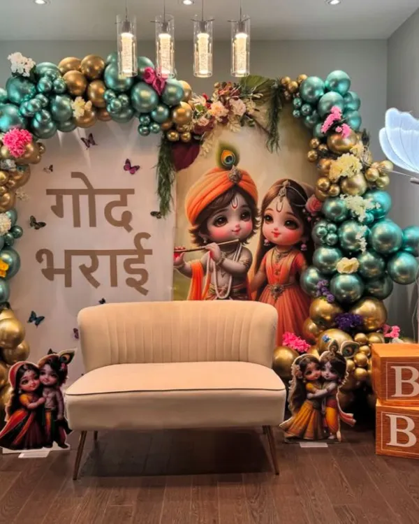 A beautiful 'Godh Bharai' ceremony with a Krishna theme. The decor features a backdrop of baby Krishna and Radha, surrounded by a teal and gold balloon arch and neon butterfly lights.