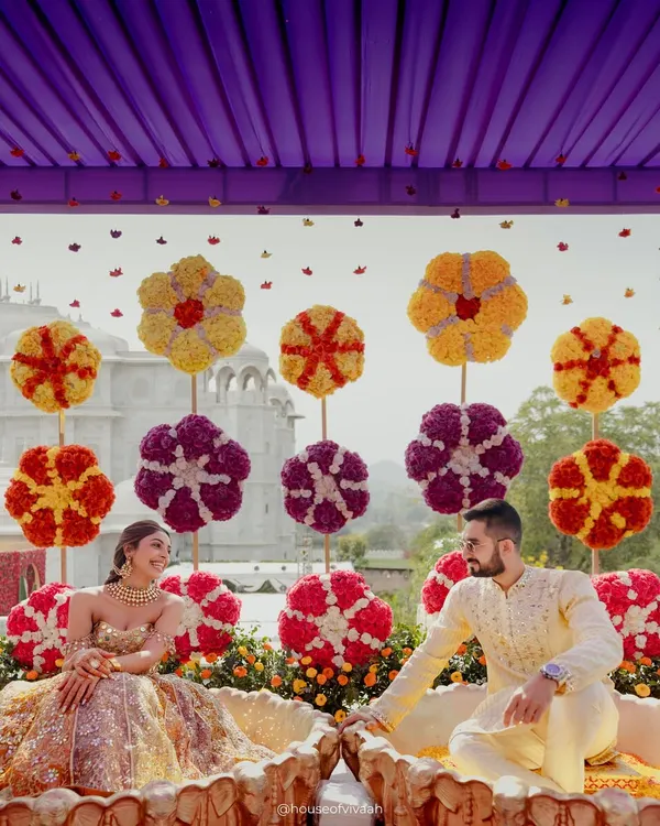 The bride and groom seated for their Haldi ceremony against a backdrop of giant, handcrafted floral lollipops. The playful design and vibrant colors under a lilac canopy create a joyful and unique setting.