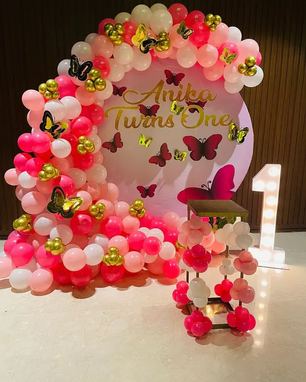 A beautiful butterfly-themed first birthday setup in shades of pink. The circular backdrop is framed by a dense balloon arch, with gold and pink butterfly stickers adding detail throughout the design.