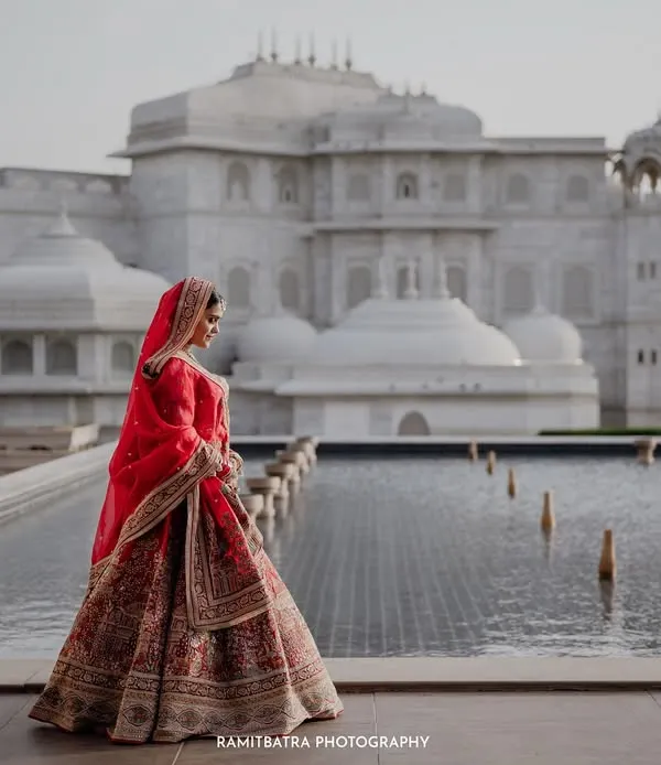 A bride in her stunning red lehenga stands against the magnificent architecture of Fairmont Jaipur, a portrait of regal grace.
