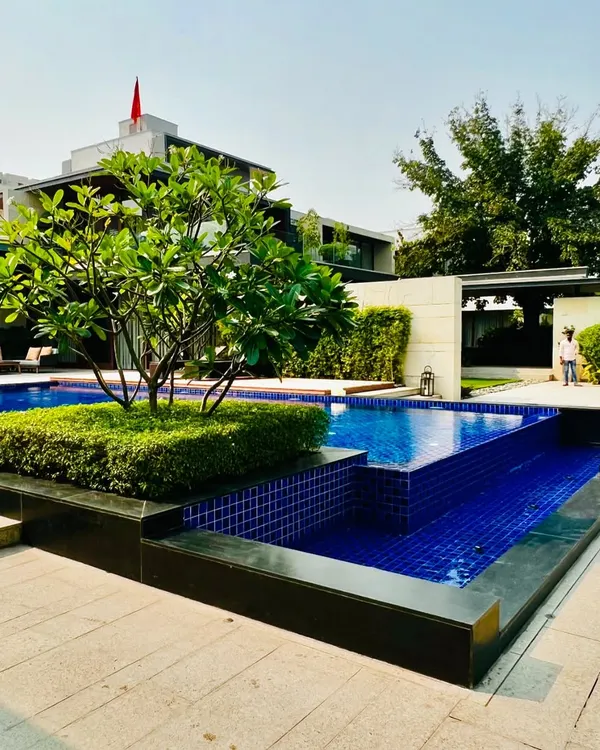 A stunning poolside at a Sultanpur farmhouse, featuring a Frangipani tree on a small island, multi-level water bodies, and a clear view of the modern home architecture.