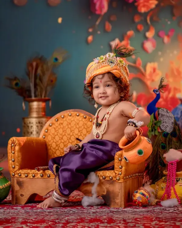 A joyful portrait of a baby boy dressed as Krishna, seated on a miniature golden throne. This Janmashtami themed photoshoot uses a rich, colorful backdrop and detailed props like a small matki to create a divine and memorable image.