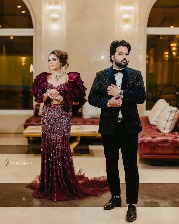 A stylish, full-length portrait of the couple at their reception, showcasing the bride's dramatic ruffled gown and the groom's classic tuxedo.