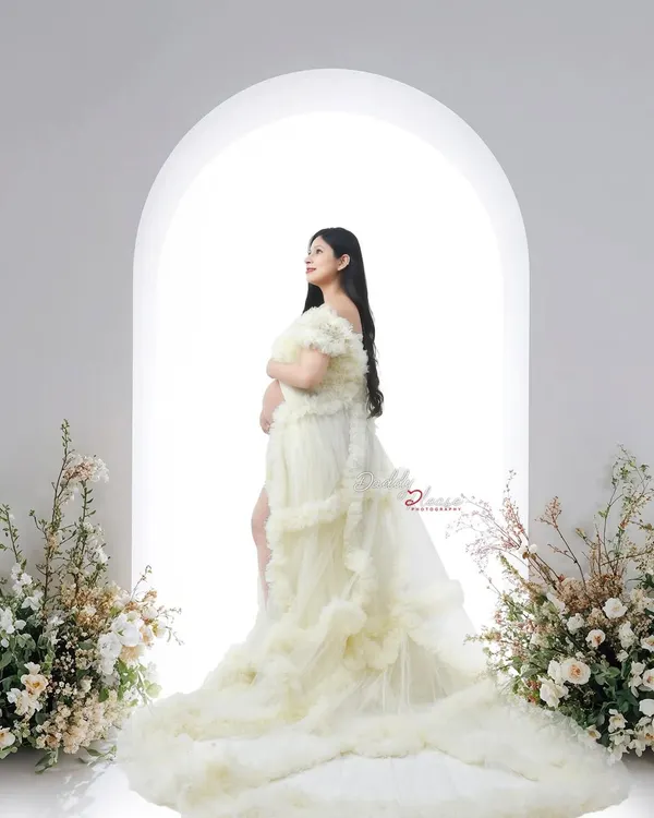 A beautiful silhouette of an expectant mother in a cream-colored ruffled gown. The bright light from the arched window behind her creates a heavenly, glowing effect.