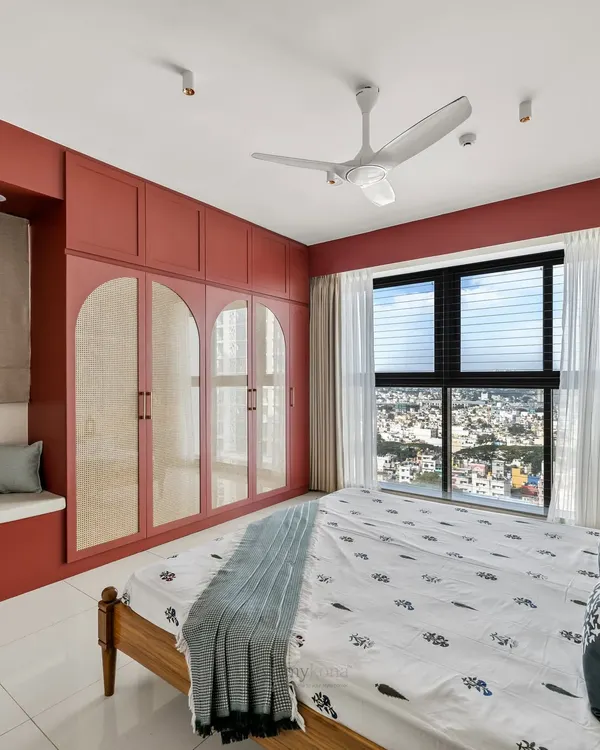 This master bedroom in a Shapoorji Pallonji flat features a bold burgundy red wardrobe with arched cane detailing. It proves that a strong color choice can create a warm, inviting, and highly functional space, complete with a cozy bay window seat.