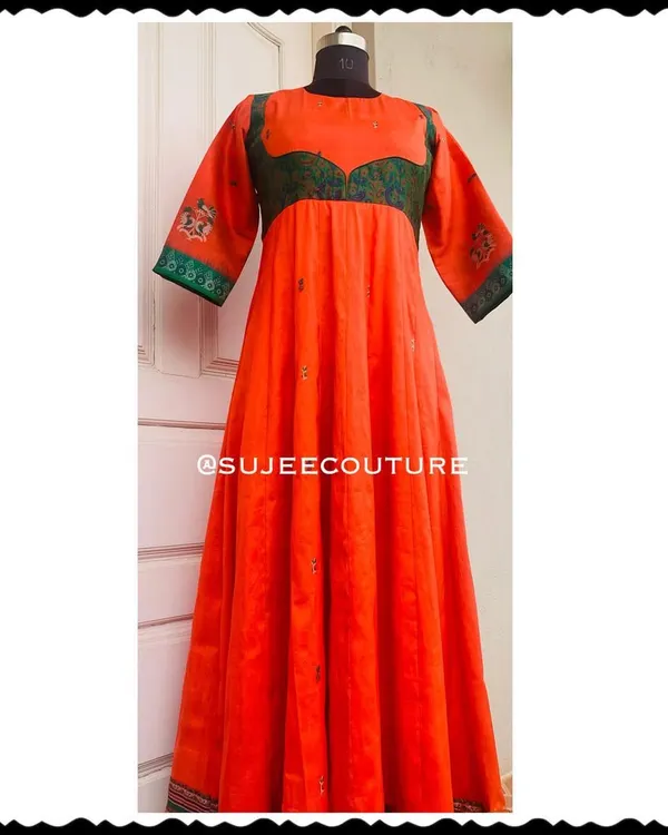 A full-length view of a custom Anarkali-style gown. This vibrant orange dress features a contrasting green yoke made from brocade fabric, with subtle embroidery on the sleeves and hem.
