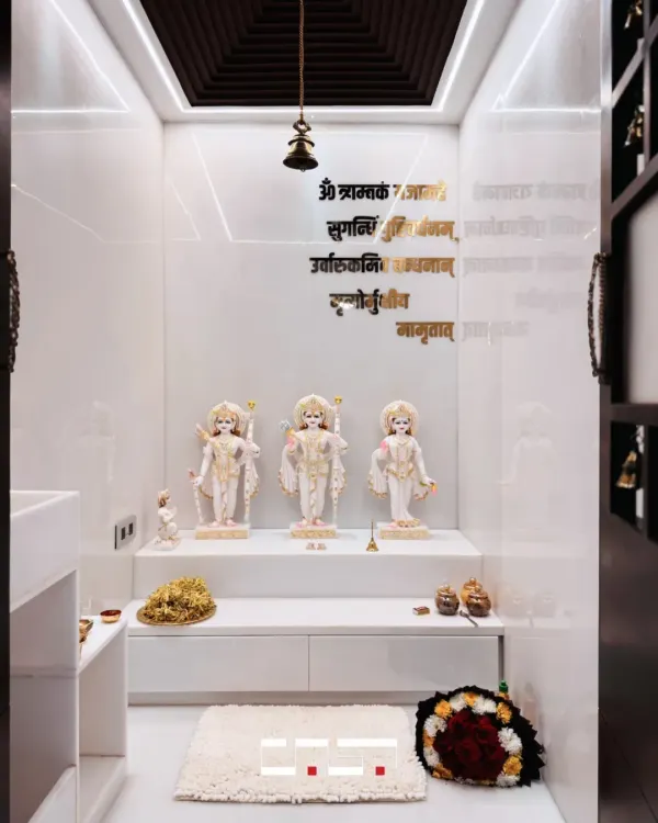This minimalist home mandir creates a sanctuary of peace. The design features pristine white marble idols, a clean white platform with storage, and the sacred Gayatri Mantra in elegant gold lettering on the wall.
