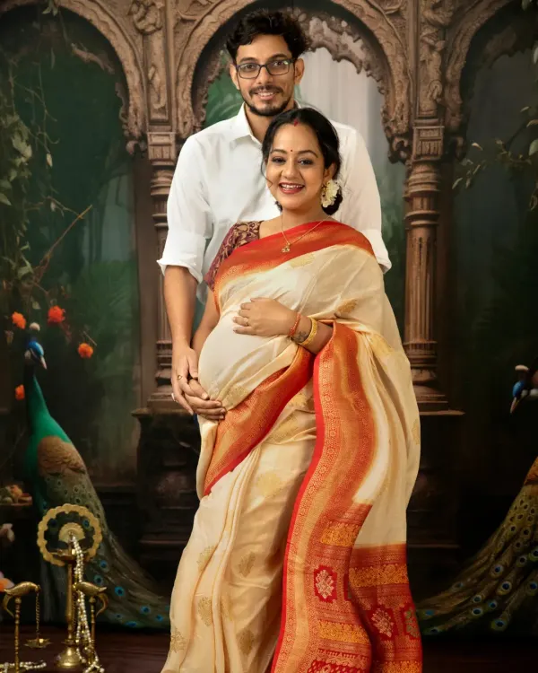 A classic couple portrait from a traditional maternity session. The mother-to-be is radiant in a cream and red silk saree, standing with her partner against a backdrop inspired by Indian art, complete with peacocks.